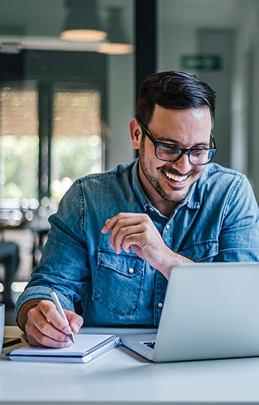 Man with glasses smiling while using his computer and taking notes