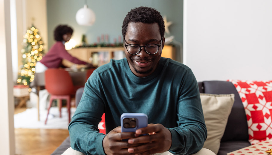 Man looking at his phone smiling with woman and christmas tree in background out of focus