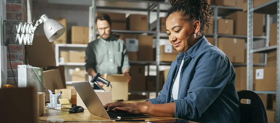 Woman on her computer in her small business with a man in the background holding a box