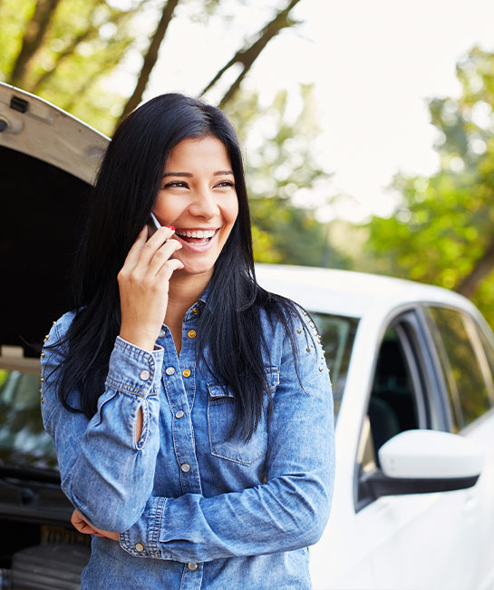 Woman leaning against her broken down car smiling while speaking on her phone